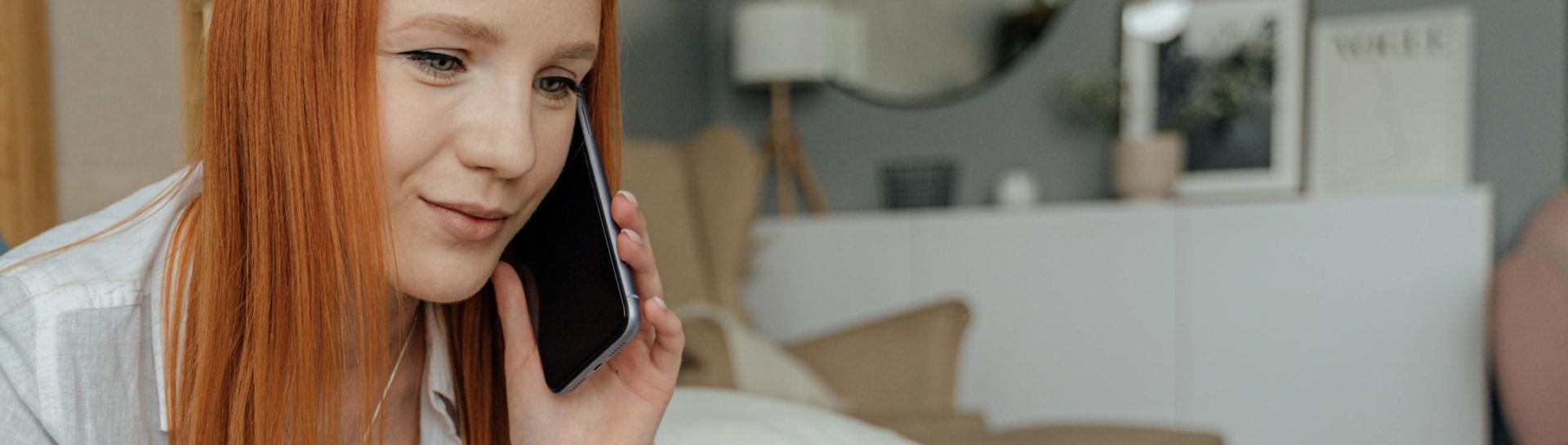 Red-haired woman multitasking in a cozy bedroom, using a laptop and phone for work.