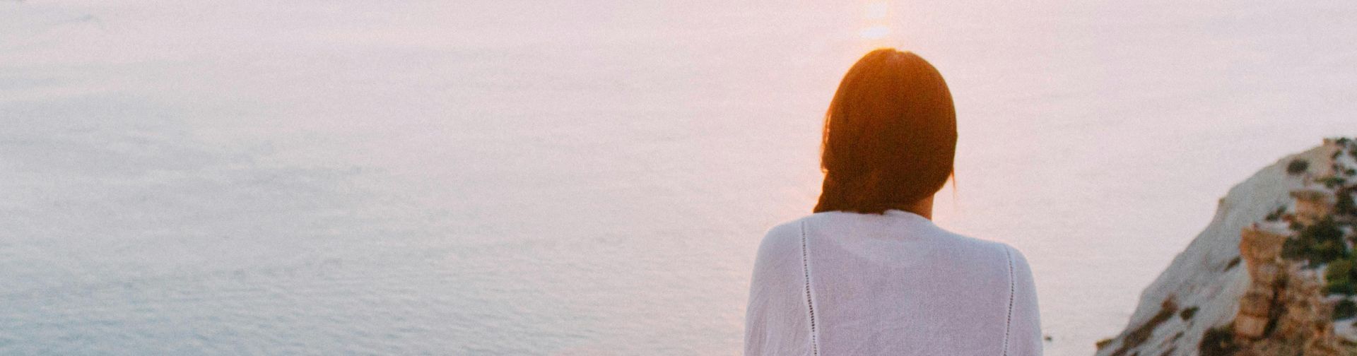 A serene view of a woman sitting on a rocky cliff, gazing at the ocean during sunset. Perfect for relaxation and travel themes.